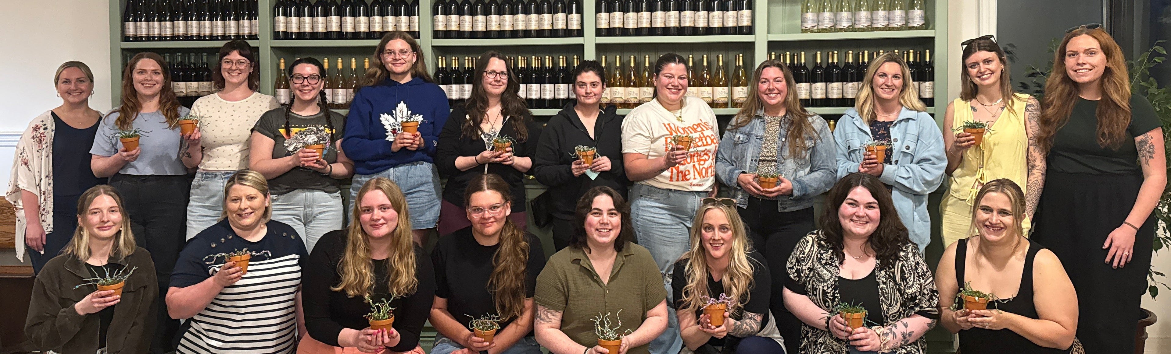 Group of people posing with their crafts at CRAFT CLUB in front of a wall of bottles in a store.
